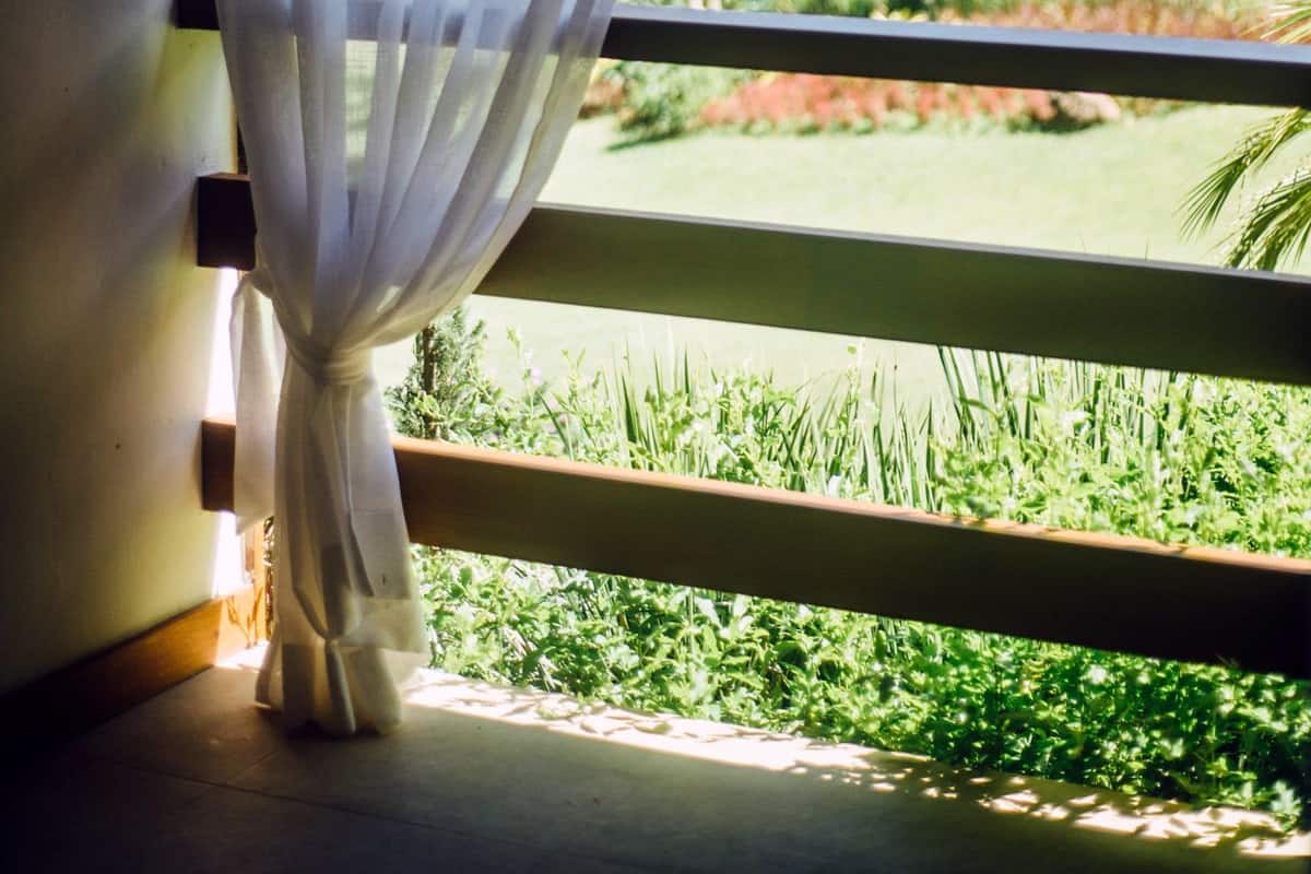 Une fenêtre ornée de rideaux blancs et légers, une balustrade en bois et une vue sur un jardin luxuriant, où poussent différentes plantes et arbres.
