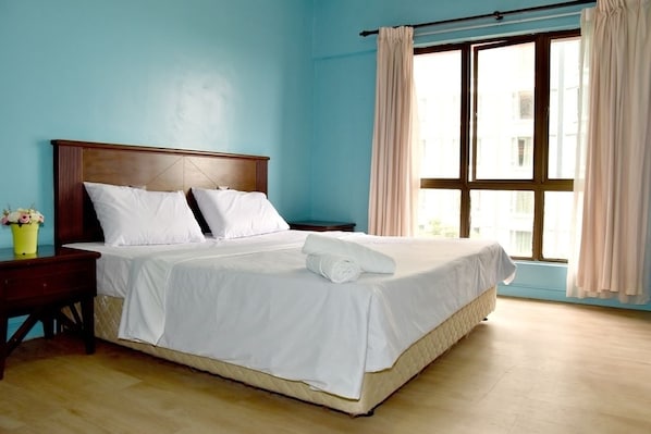 A neatly made bed with white linens and a rolled towel, a wooden nightstand with a vase of flowers, and a window with curtains.