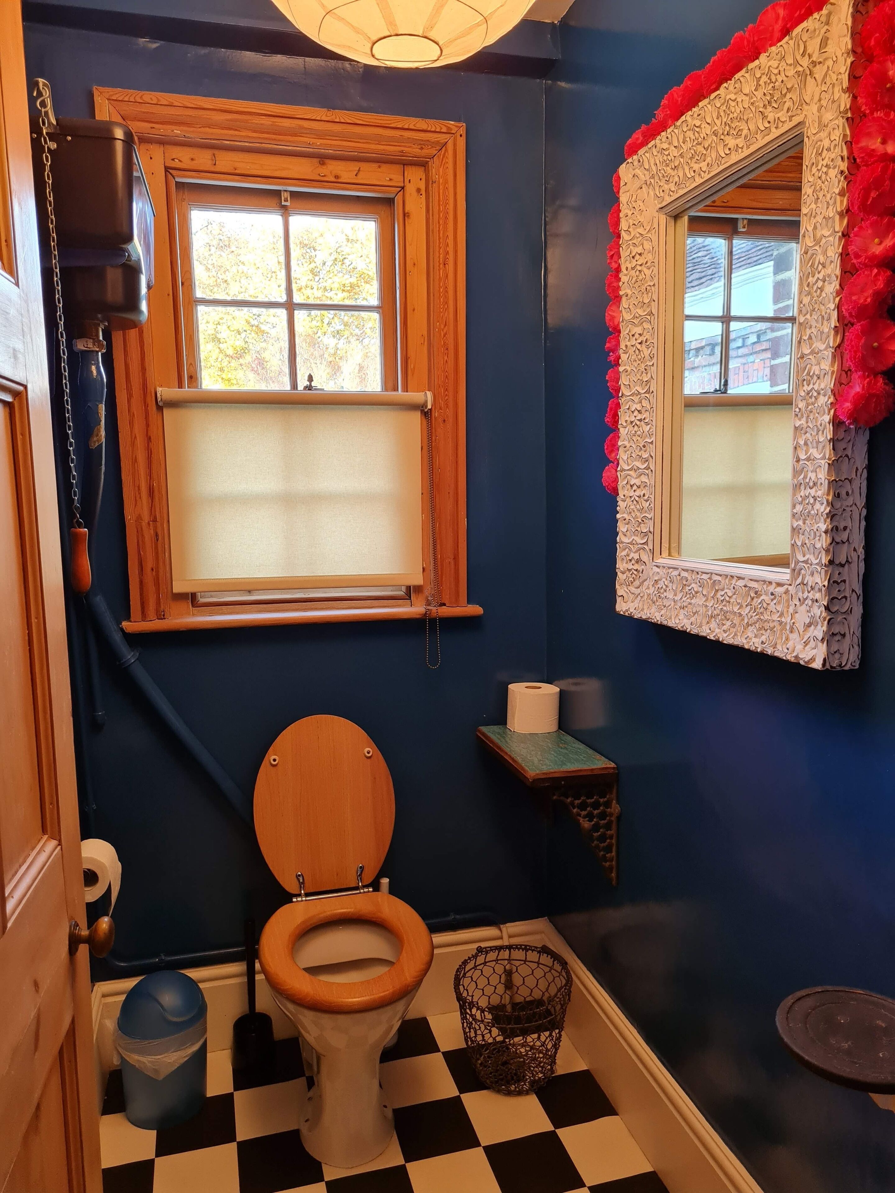 Ein Badezimmer mit einer Holzwc, einem Fenster mit Holzrahmen und einem dekorativen Spiegel mit roten Blumen.