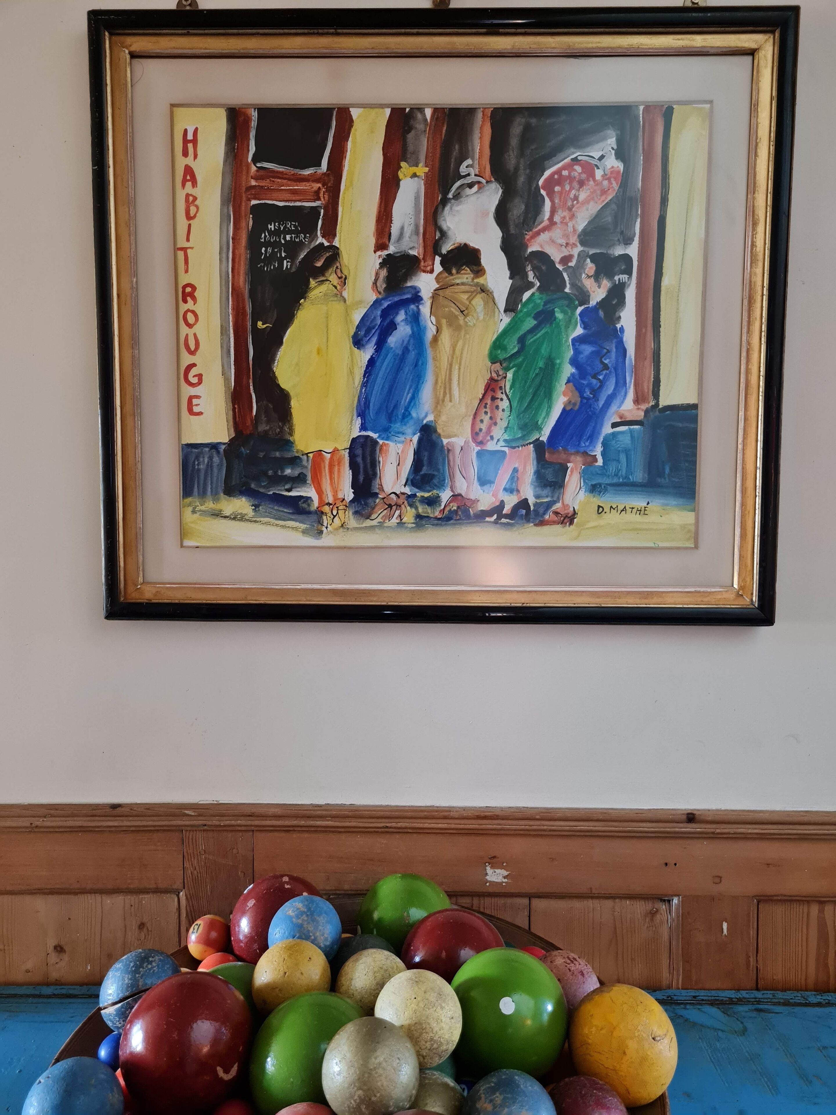 Een lijstwerk schilderij met de tekst 'HAFTROGE' en 'D. MATHE' op een muur. Daaronder een houten kastje met kleurrijke bowlingballen.
