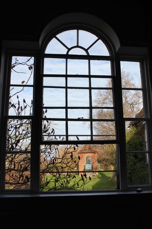 A large arched window with multiple panes, offering a view of a brick building and surrounding trees. - St John's Croft (Winchester)