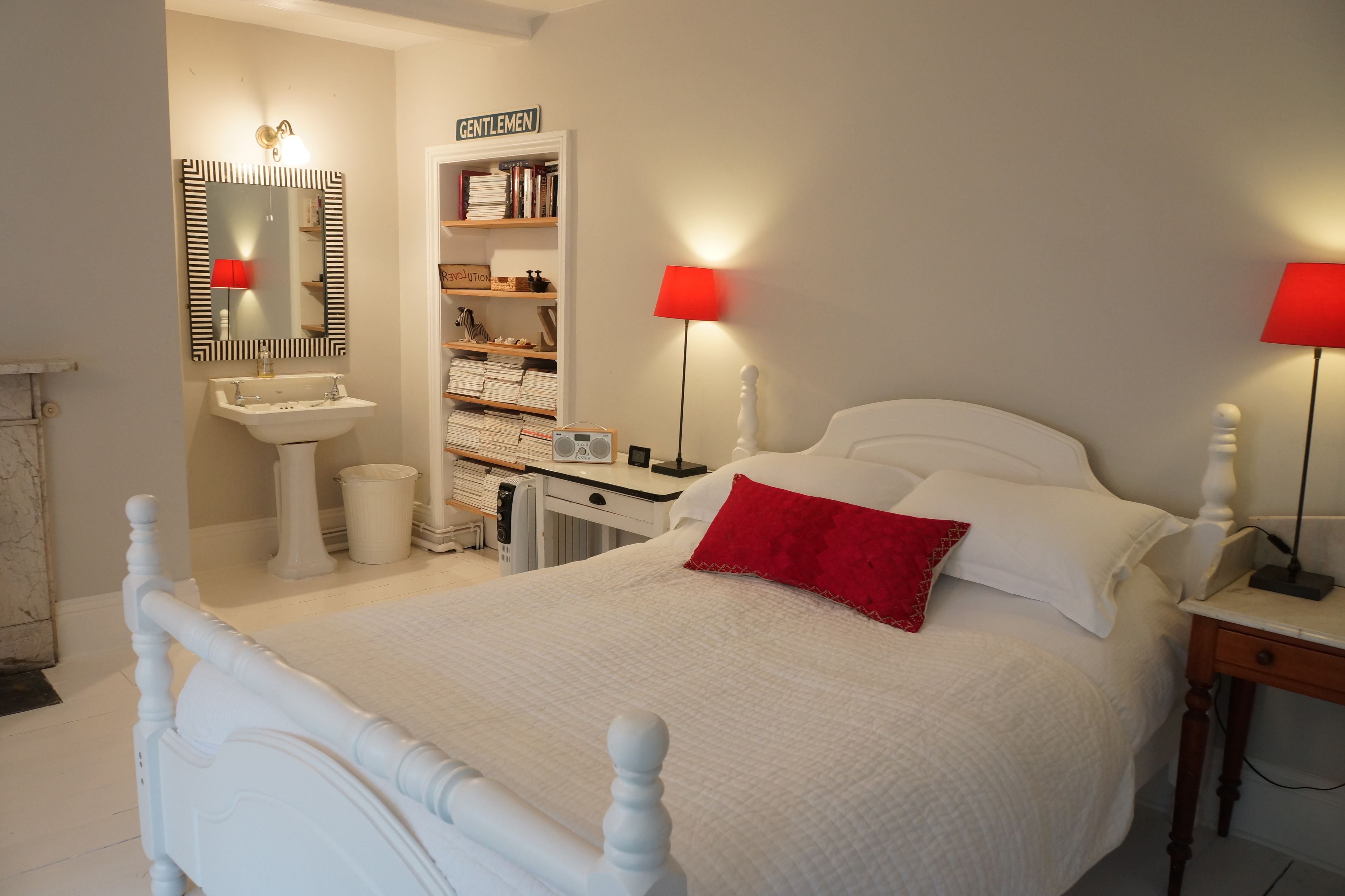 A bedroom with a white bed, red pillows, a bedside table, a nightstand, a bookshelf, and a mirror.