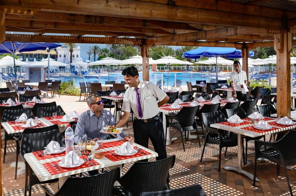 Un área de comedor al aire libre con mesas preparadas para dos personas, una piscina al fondo y sombrillas que dan sombra.