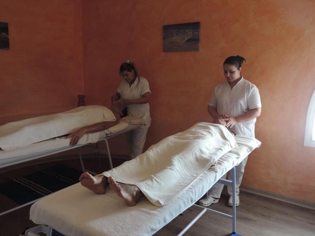 A room with two beds, one occupied by a person lying on a bed, and another person standing beside it.