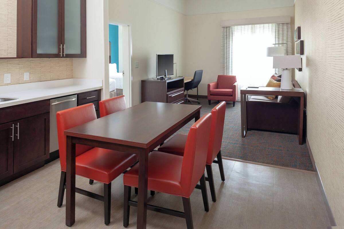A hotel room with a dining area, a TV, a desk with a lamp, a red chair, and a kitchen area with cabinets and a sink.