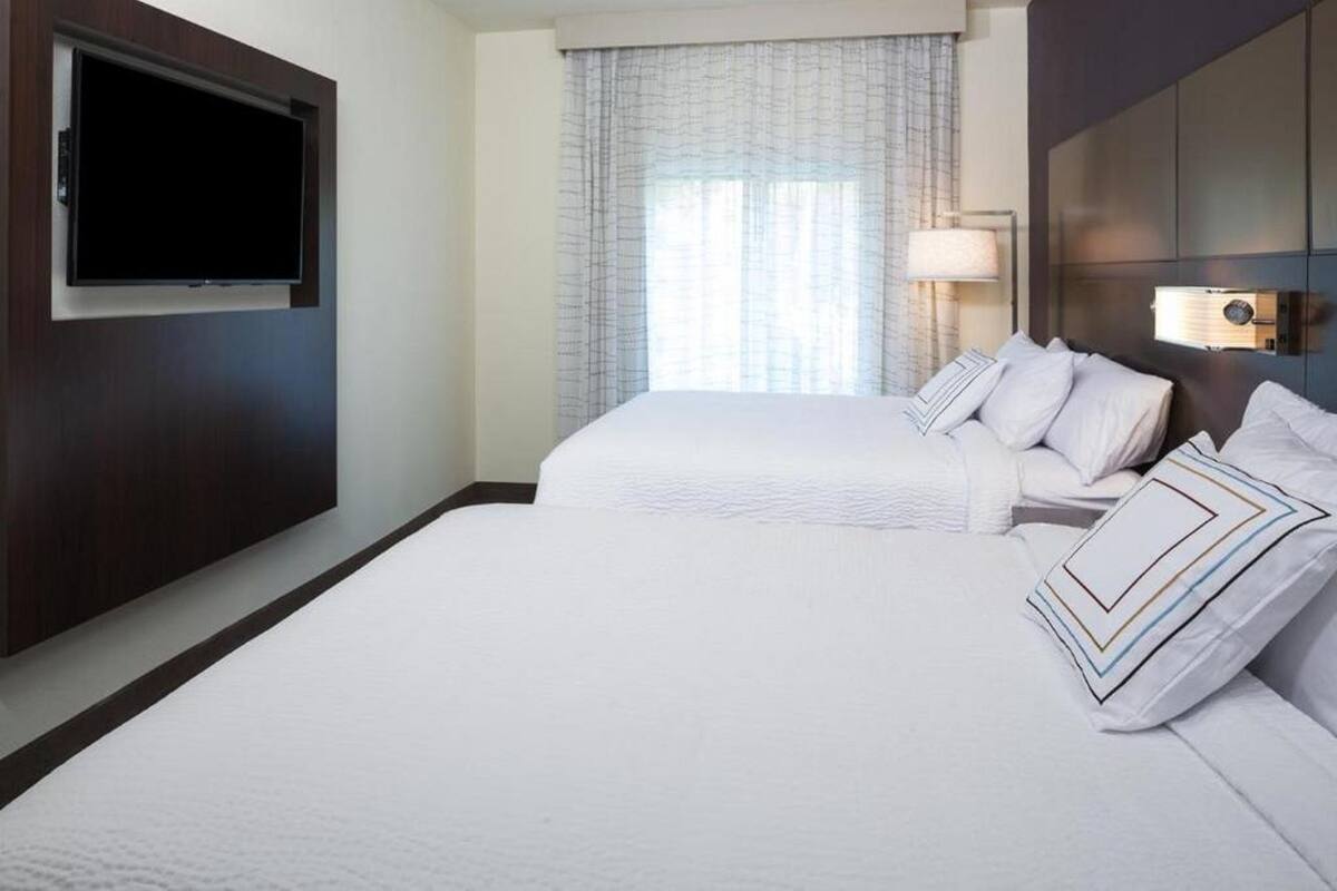 A hotel room with two beds, a flat-screen TV, a window with curtains, and a bedside lamp.