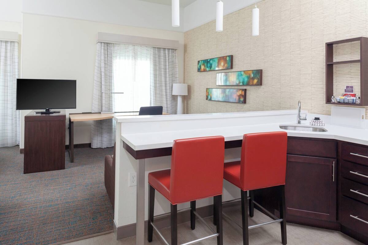 A hotel room with a kitchenette, a flat-screen TV, a desk, two red chairs, a large window with curtains, and artwork on the wall.