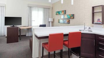Habitación de hotel con kitchenette, una televisión de pantalla plana, escritorio, dos sillas rojas, un ventanal con cortinas y obras de arte en la pared.