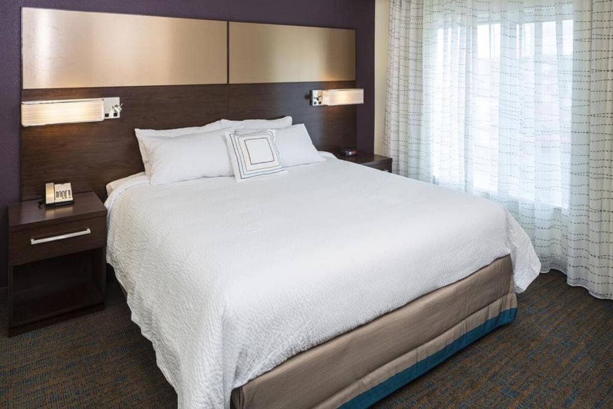 A hotel room with a large bed, a headboard with built-in lighting, a bedside table with a phone, and a window with sheer curtains.