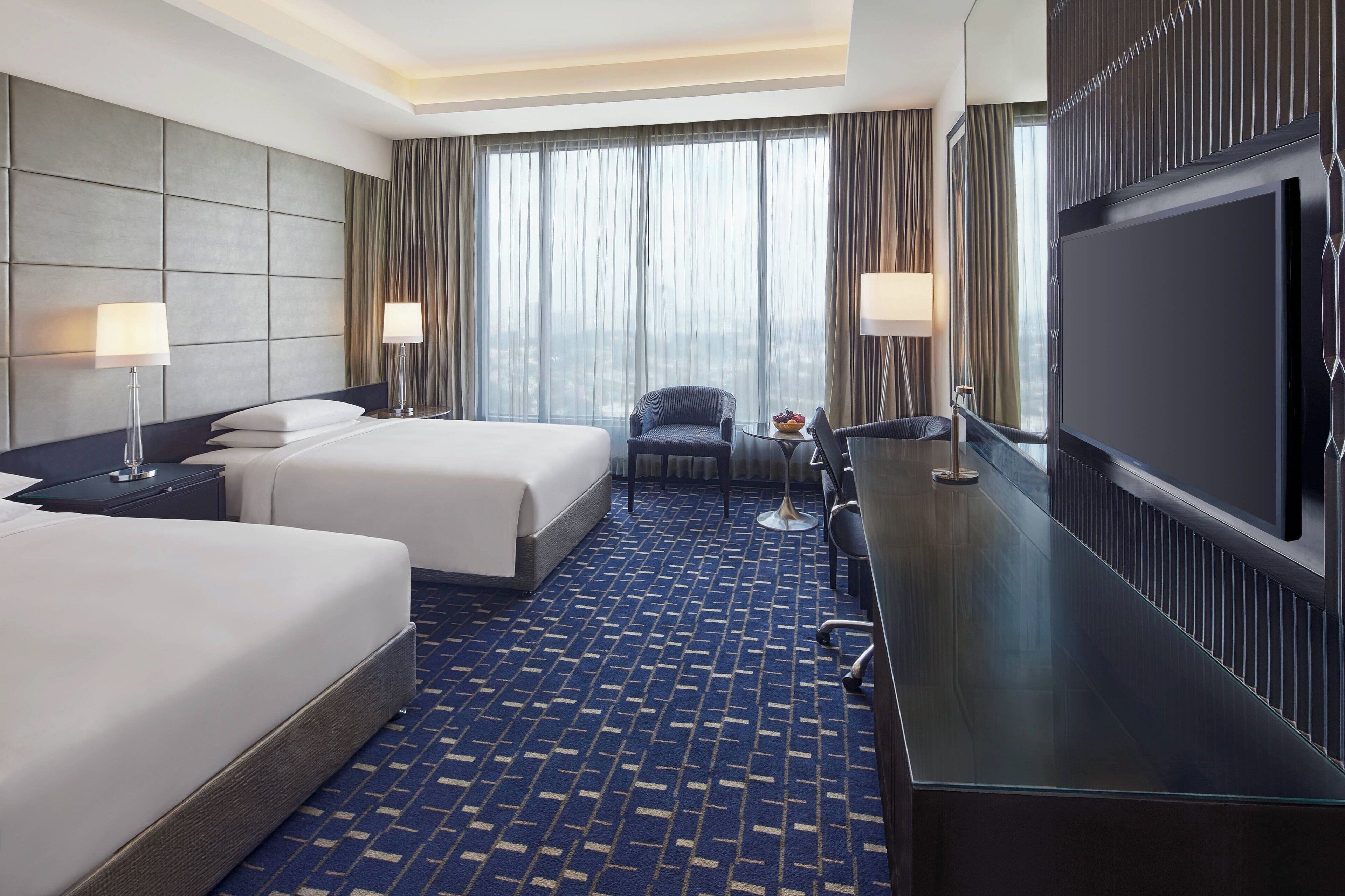 A hotel room with two beds, a desk with a TV, a chair, and a large window with curtains.