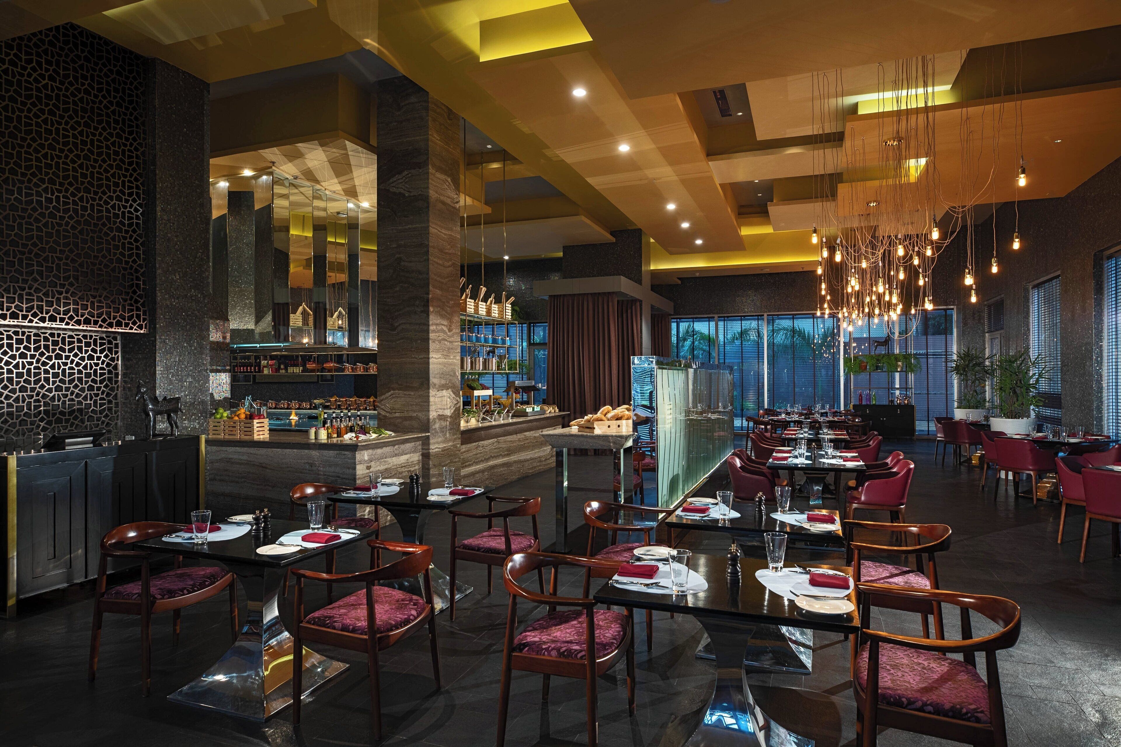 Un restaurante moderno con techos altos, lámparas colgantes y una zona de bar. Las mesas están dispuestas con sillas tapizadas en rojo y cristalería.