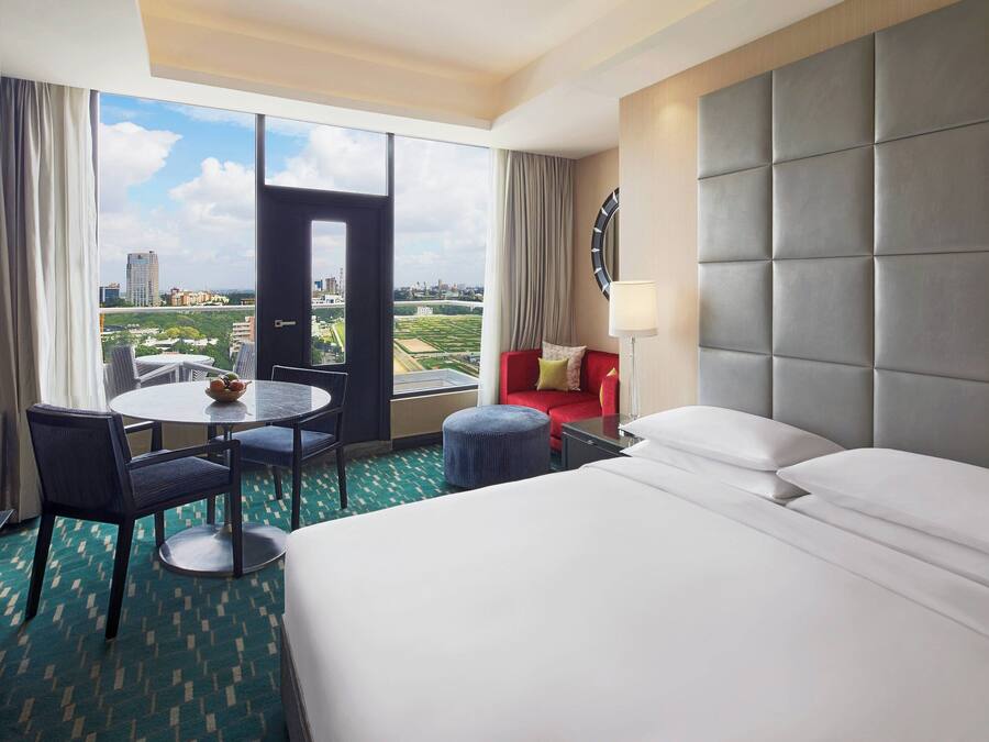 A hotel room with a large bed, a round dining table, two chairs, a red sofa, and a view of the cityscape through large windows.