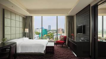 A hotel room with a large bed, a desk, a chair, a TV, and a view of the cityscape.