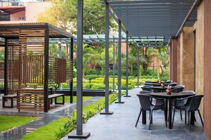 Relax in a serene garden setting with a covered patio, tables, and chairs, surrounded by lush greenery and a soothing water feature.