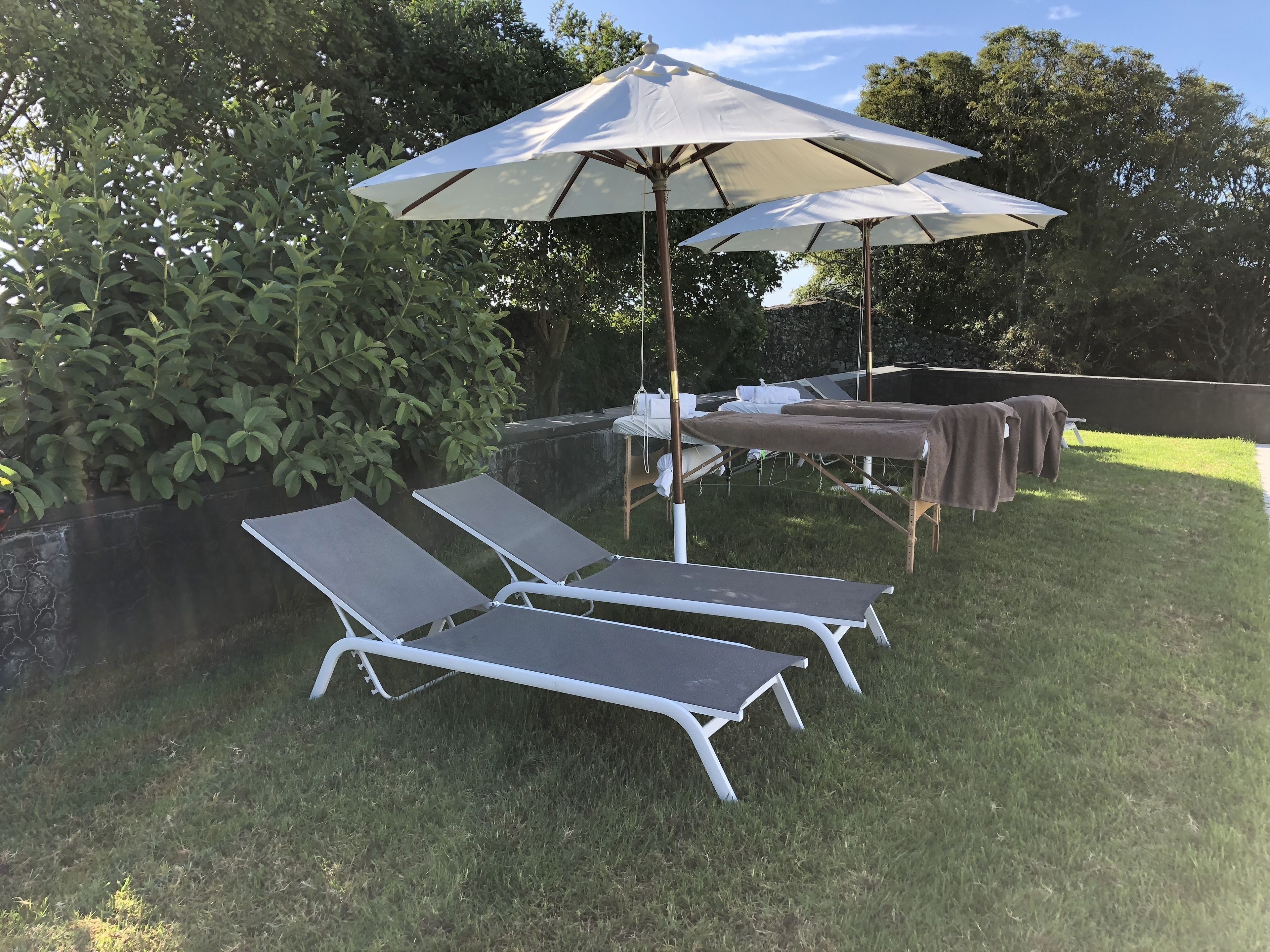 A patio area with two lounge chairs and a large umbrella, surrounded by greenery and a stone wall.