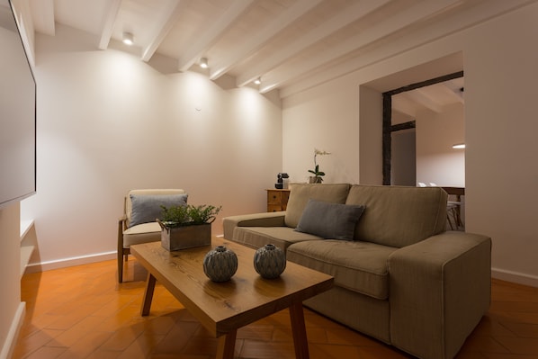 A modern living room with a sofa, a wooden coffee table, and a chair. There is a small side table with a plant and a vase. The room has wooden flooring and white walls. - Quinta de Santa Clara (Ponta Delgada)