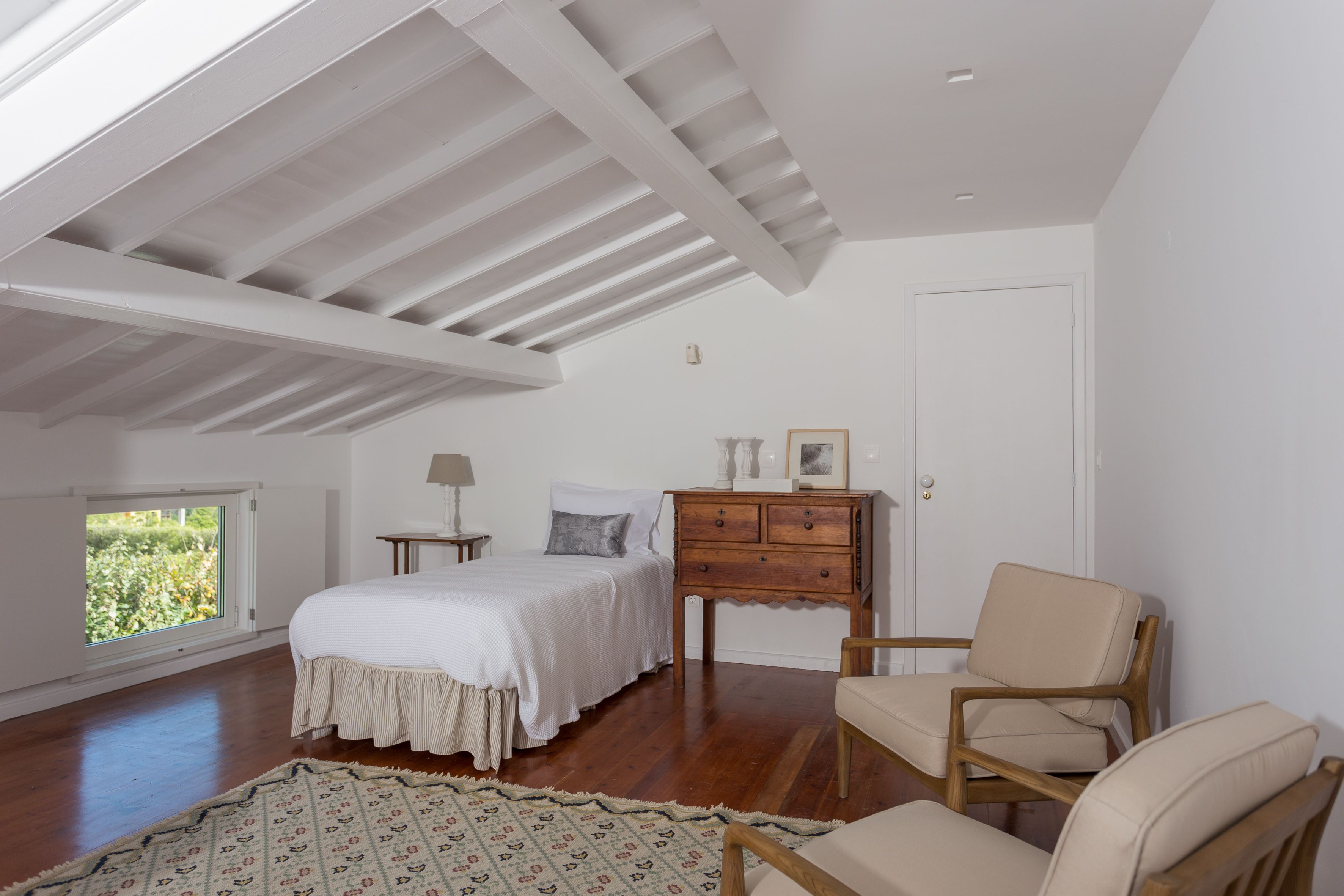 Een slaapkamer met een schuine kap, een bed, een dressoir, een stoel en een raam met uitzicht op groen.
