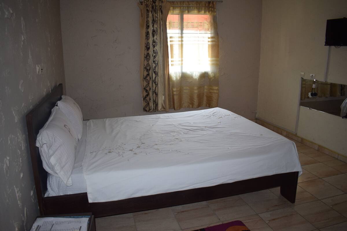 A bedroom with a bed, a window with curtains, a television mounted on the wall, and a mirror.