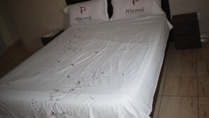 A neatly made bed with white bedding and floral patterns, two pillows with the logo 'PERONNE', and a nightstand with a phone and a small drawer. - Hôtel Résidence Péronne (Abidjan)