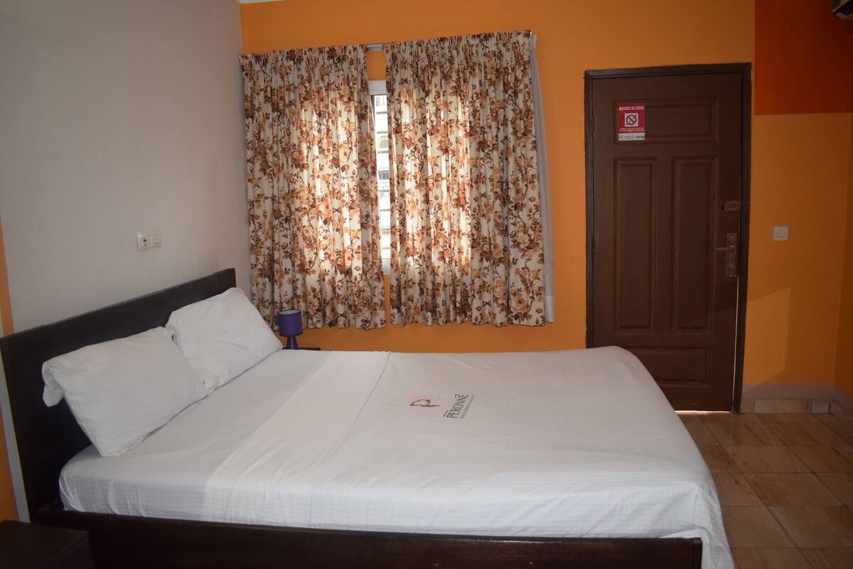 A neatly made bed with white linens, a floral curtain, and a closed door with a sign.