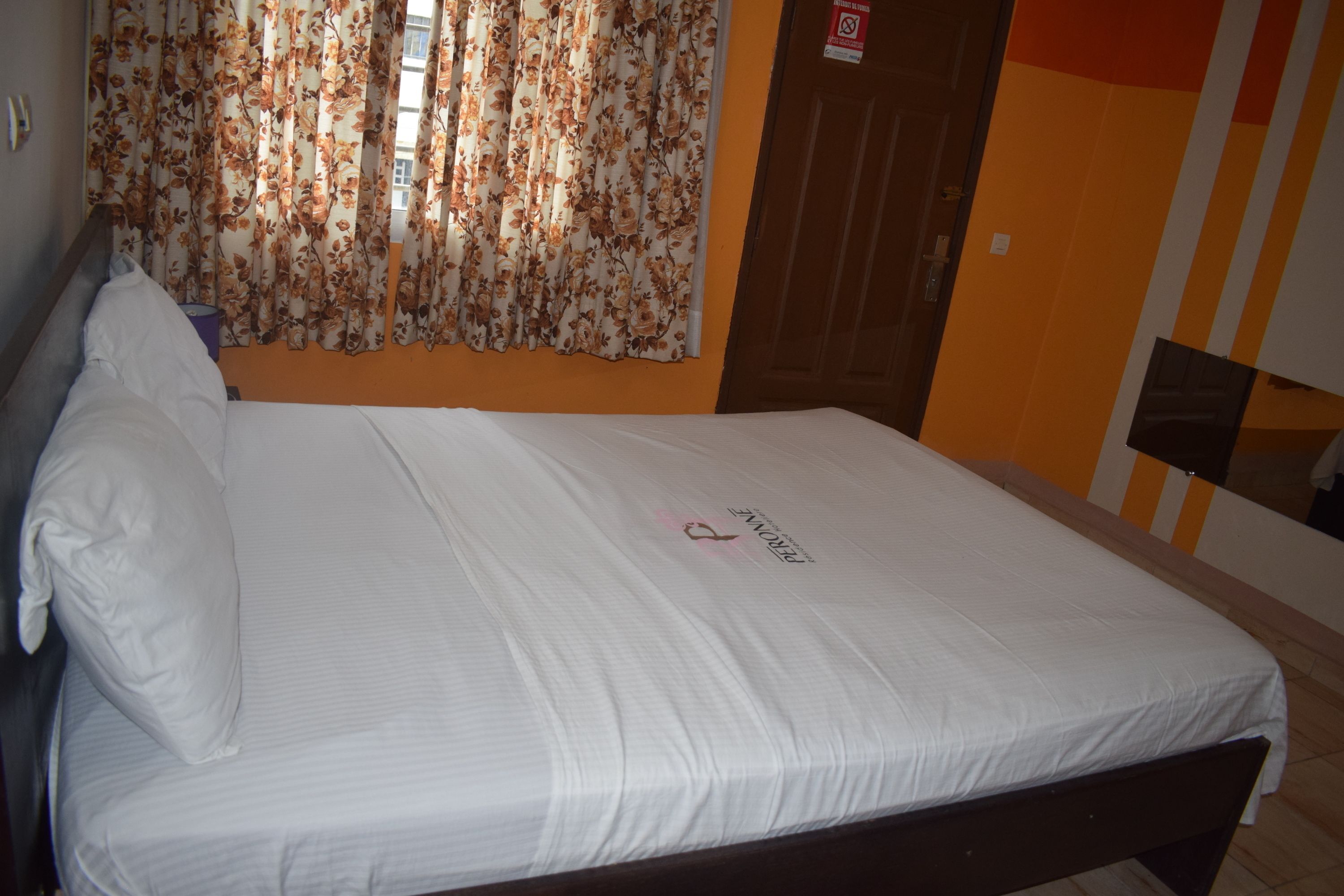 A neatly made bed with a white sheet and a decorative pillow, a window with floral curtains, and a door with a sign.