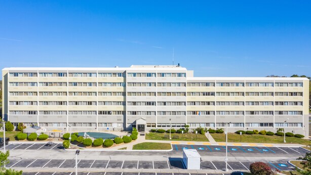 Een groot, meerlaags hotel met een centrale ingang, een parkeerplaats met aangegeven plaatsen en een klein zwembad.