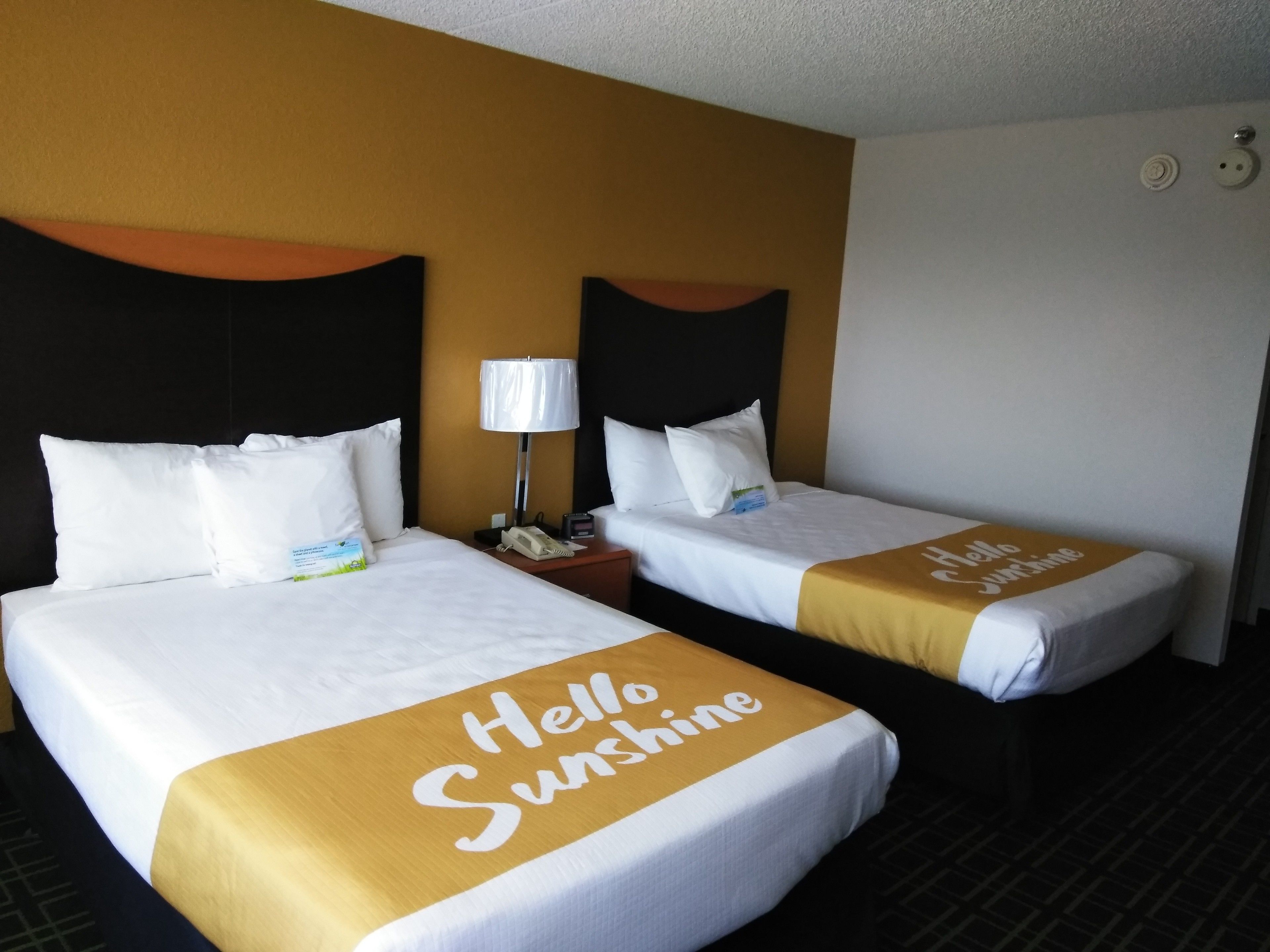 Two hotel beds with white linens and a 'Hello Sunshine' bedspread. Each bed has a black headboard and a bedside table with a lamp.