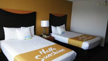 Twee hotelbedden met witte lakens en een beddegoed met de tekst 'Hello Sunshine'. Elk bed heeft een zwart hoofdbord en een nachtkastje met een lamp.