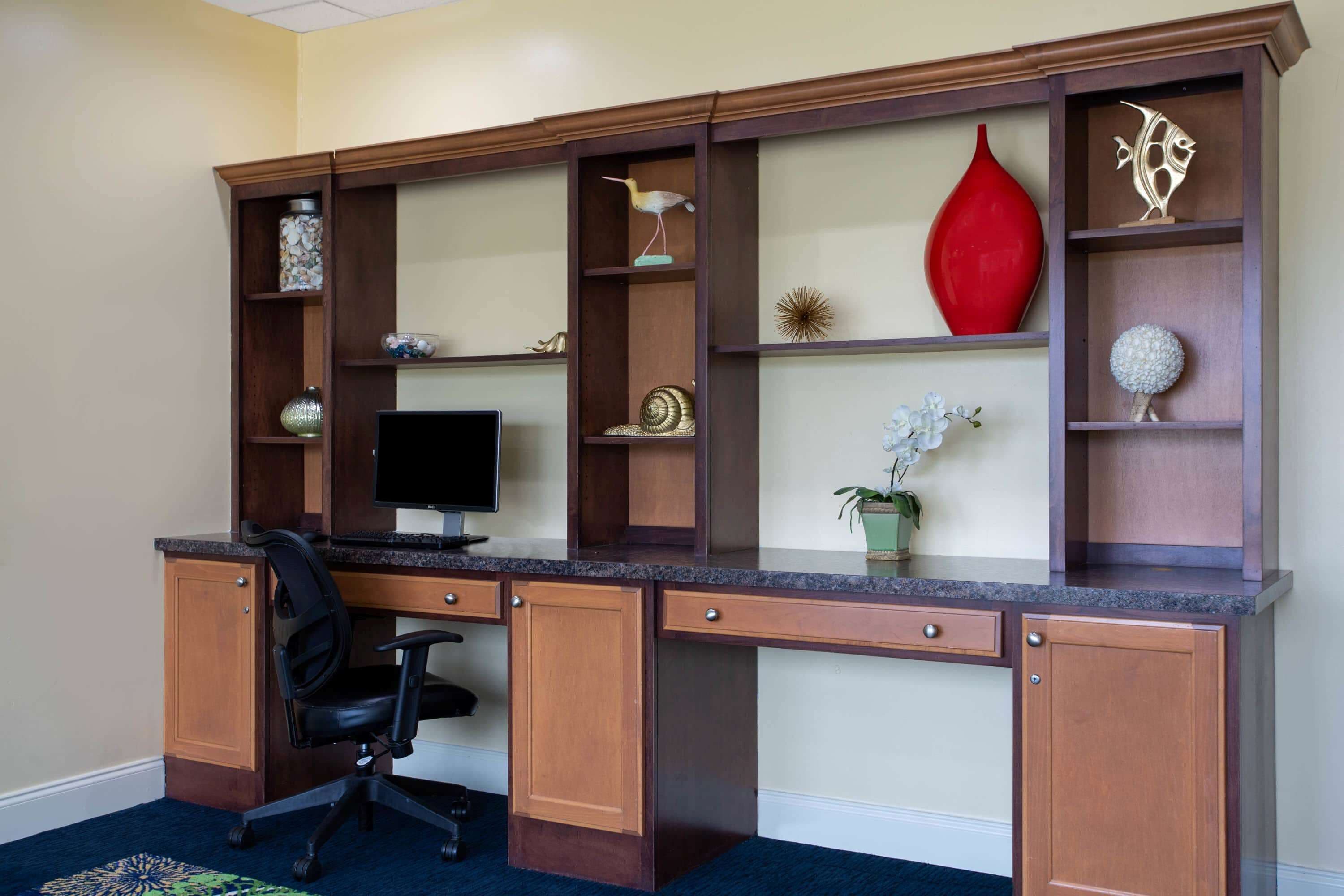 A home office with a desk, computer, and office chair. There are shelves with decorative items and a red vase.