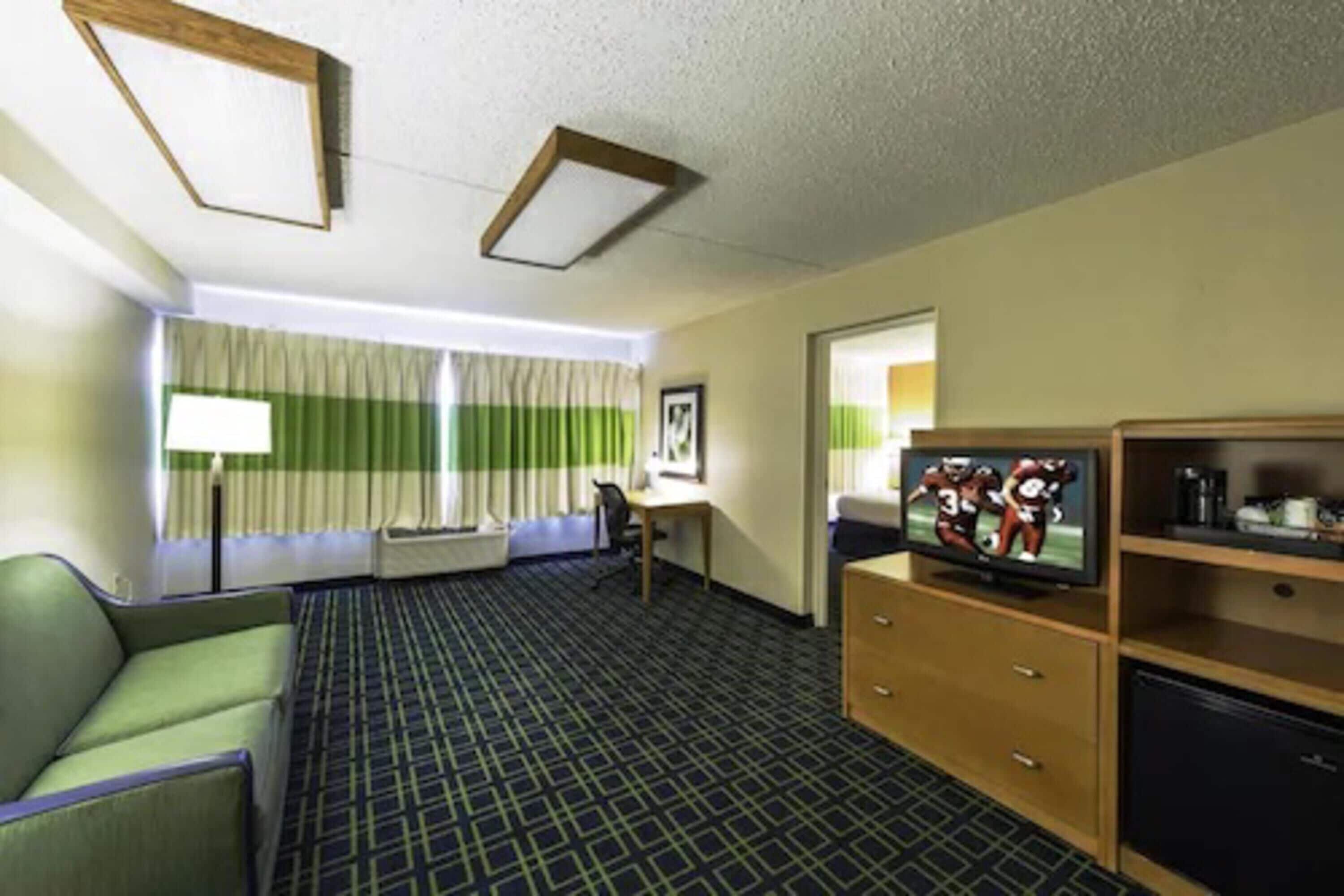 Une chambre d’hôtel avec un canapé vert, un téléviseur posé sur un meuble en bois, un bureau avec une chaise et une grande fenêtre avec des rideaux.