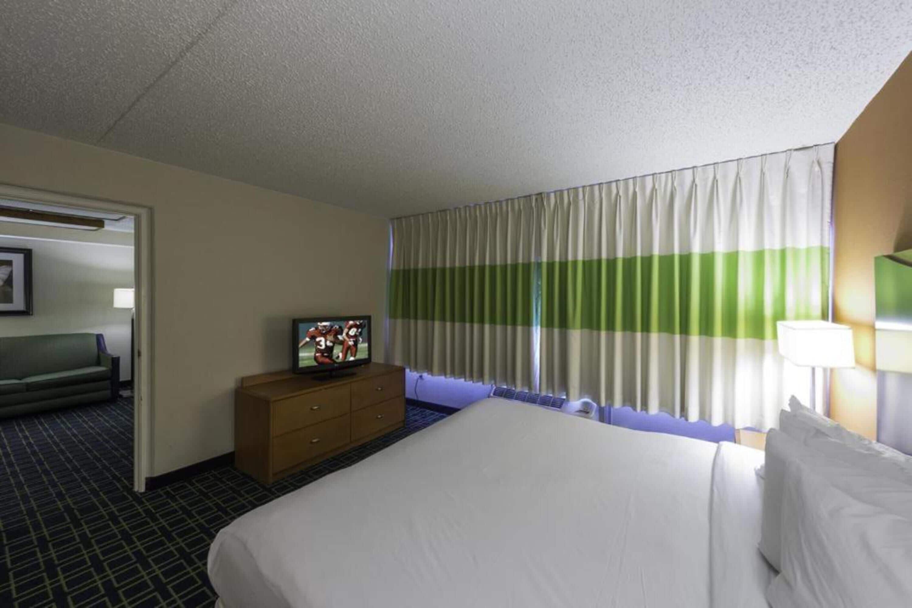 A hotel room with a bed, a TV on a dresser, a window with curtains, and a sofa in the corner.