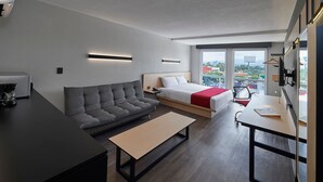 Une chambre d’hôtel moderne avec un canapé, un lit recouvert d’une couverture rouge, un bureau avec une chaise et une grande fenêtre donnant sur la ville.