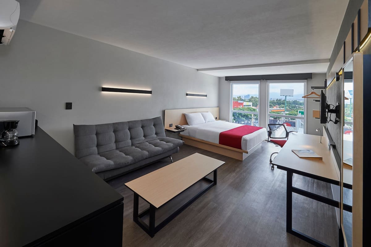A modern hotel room with a sofa, a bed with a red blanket, a desk with a chair, and a large window offering a city view.