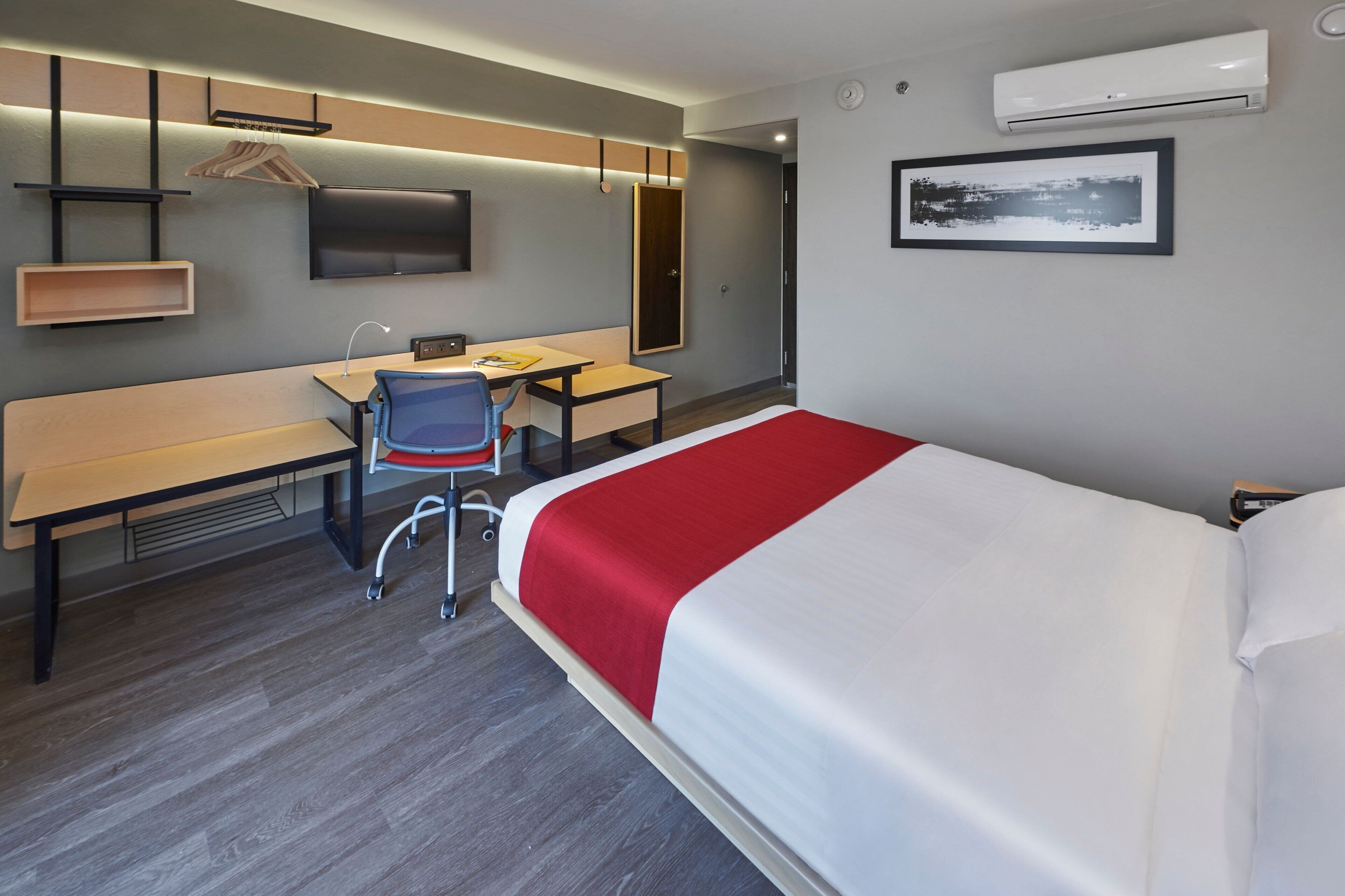 Quarto de hotel com cama, escrivaninha, cadeira e televisão fixada na parede.