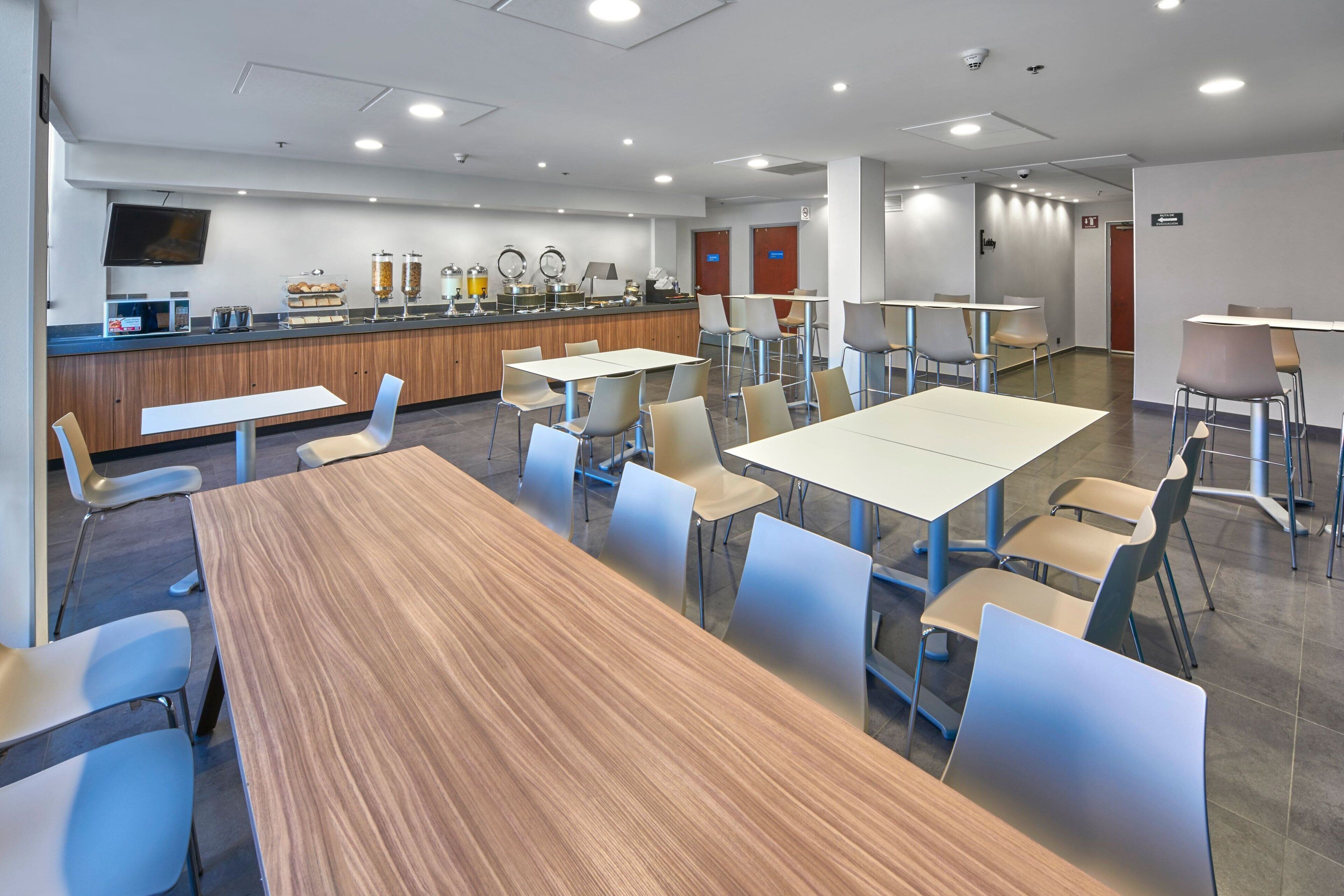 A modern dining area with a long wooden table and multiple chairs, a buffet counter with various food items, and a television mounted on the wall.