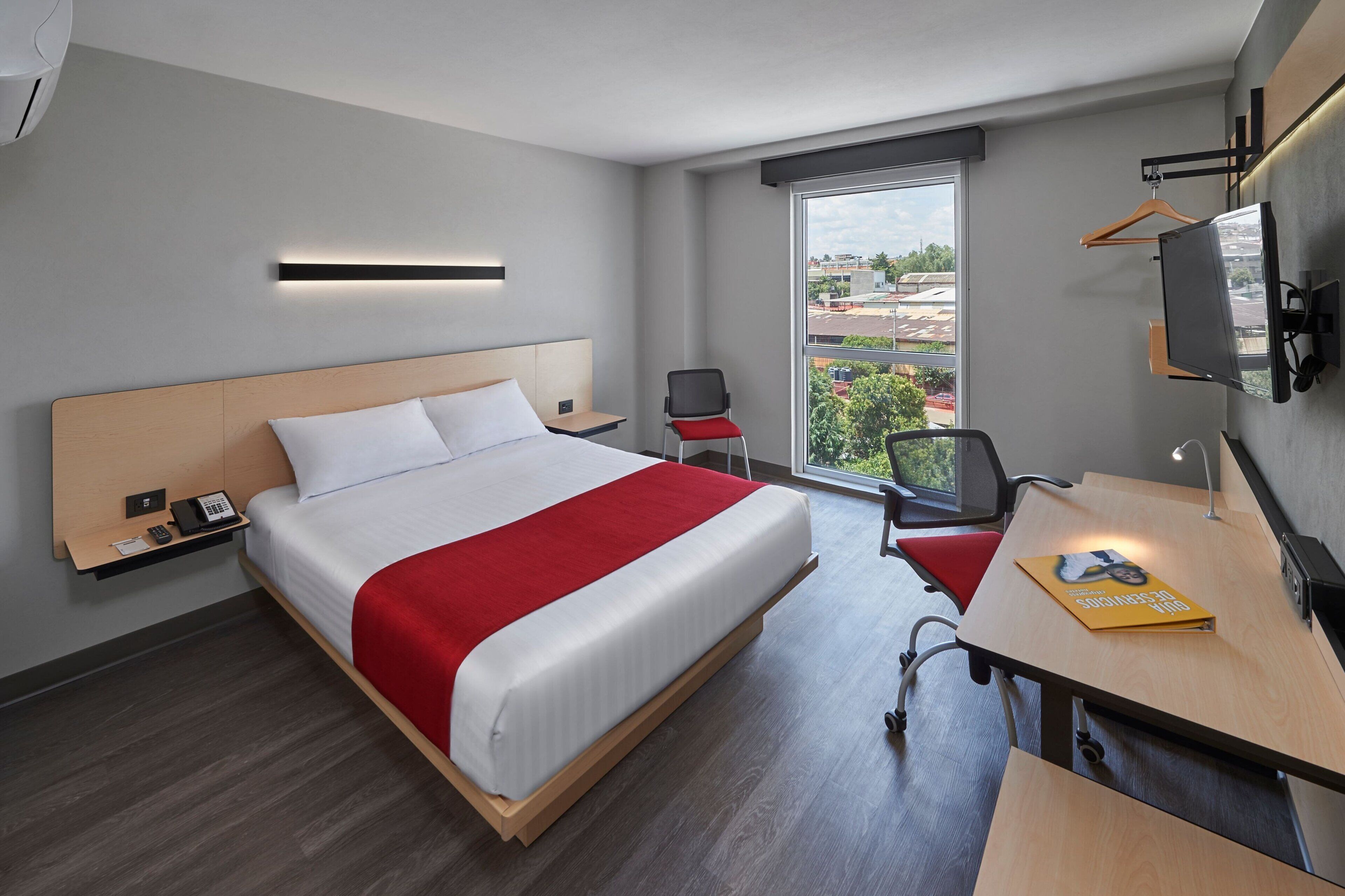 Ein modernes Hotelzimmer mit einem großen Bett, einem Schreibtisch, einem Stuhl, einem Fernseher und einem Fenster mit Blick auf die Stadt.