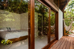 Una terraza de madera con una hamaca, una cama con vistas al bosque y una puerta de cristal que da a un balcón.