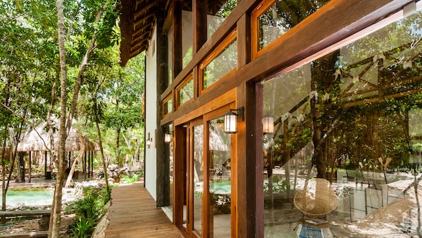 Una terraza de madera que conduce a un edificio con ventanas amplias, rodeado de árboles y una zona de piscina.