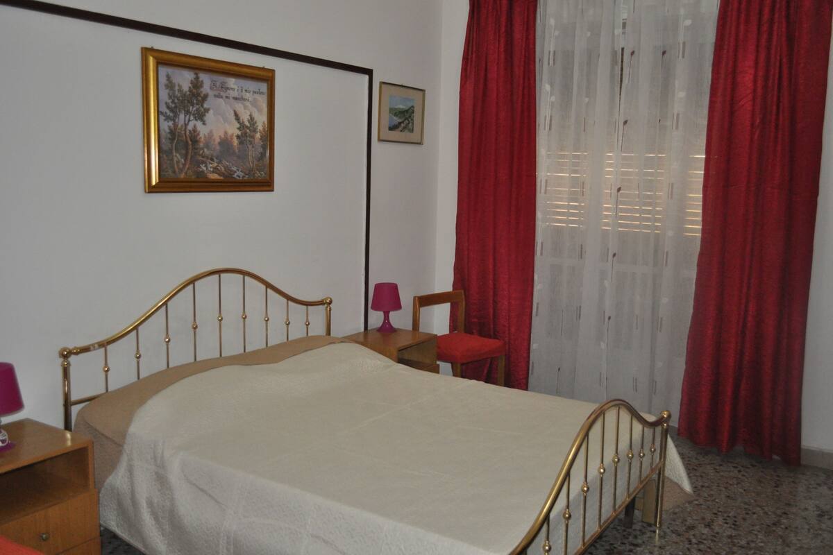 Un dormitorio con cama, mesita de noche, silla, lámpara y dos cuadros en la pared.