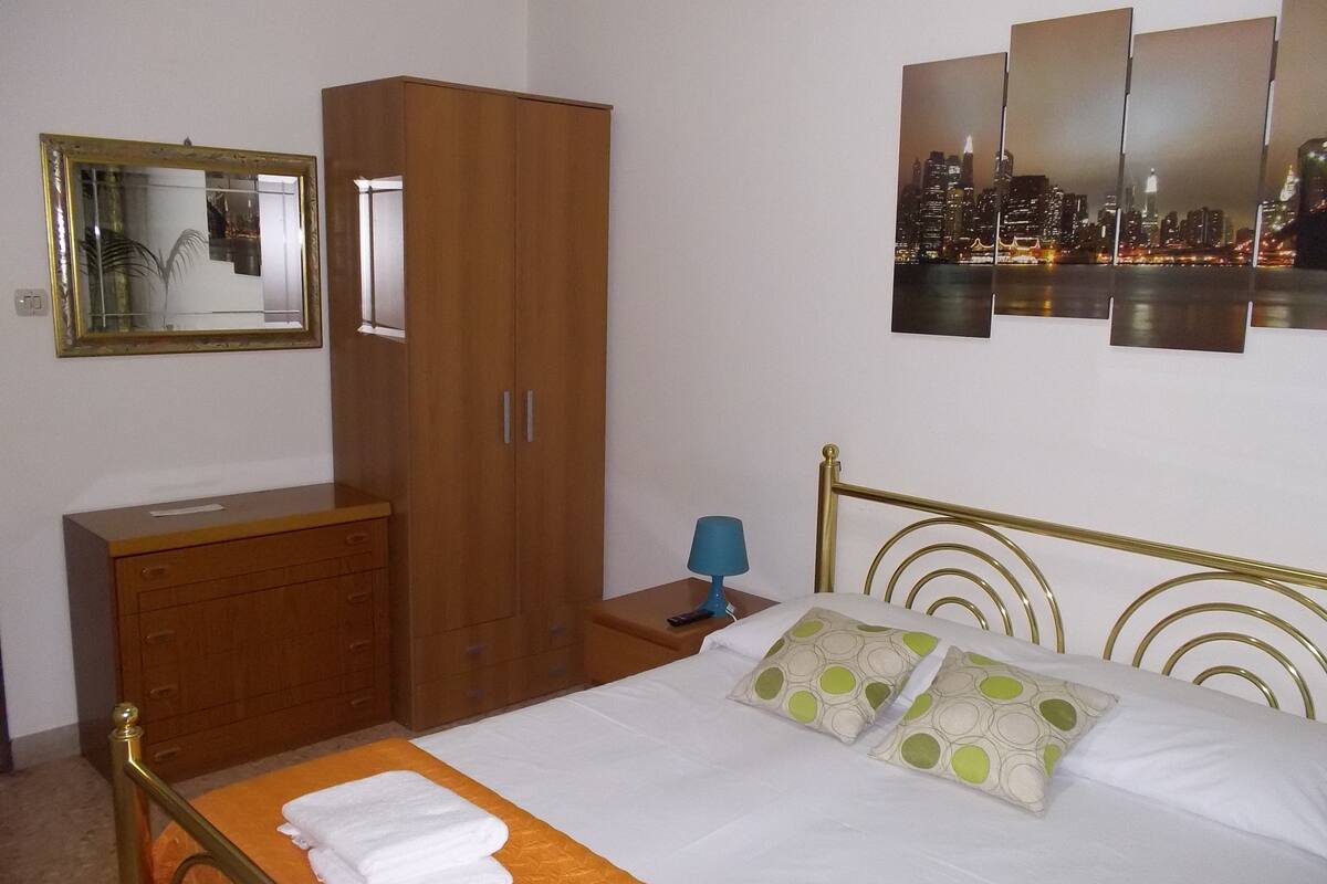 Habitación de hotel con cama, armario de madera, mesita de noche con lámpara, vista a la ciudad y espejo en la pared.