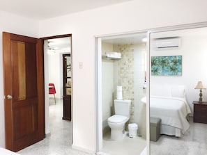A hotel room with a bathroom, a bed, a nightstand, and a wall-mounted air conditioner.