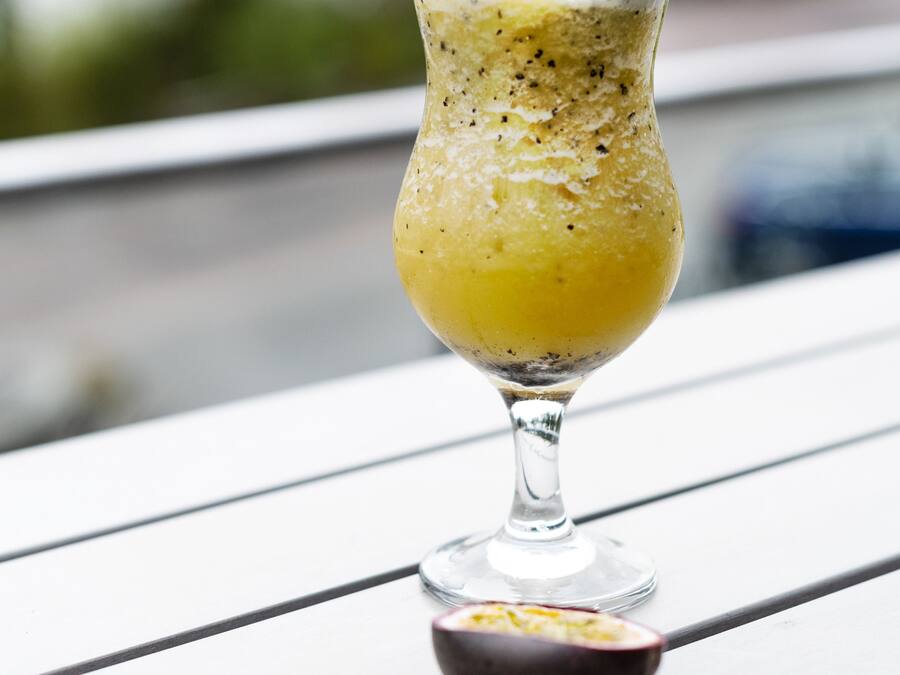A glass of yellow smoothie with a passion fruit slice and straws on a white table.