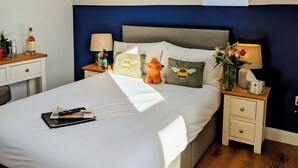 A bedroom with a bed, a nightstand, a lamp, a window, and a small table with a bottle and glasses.