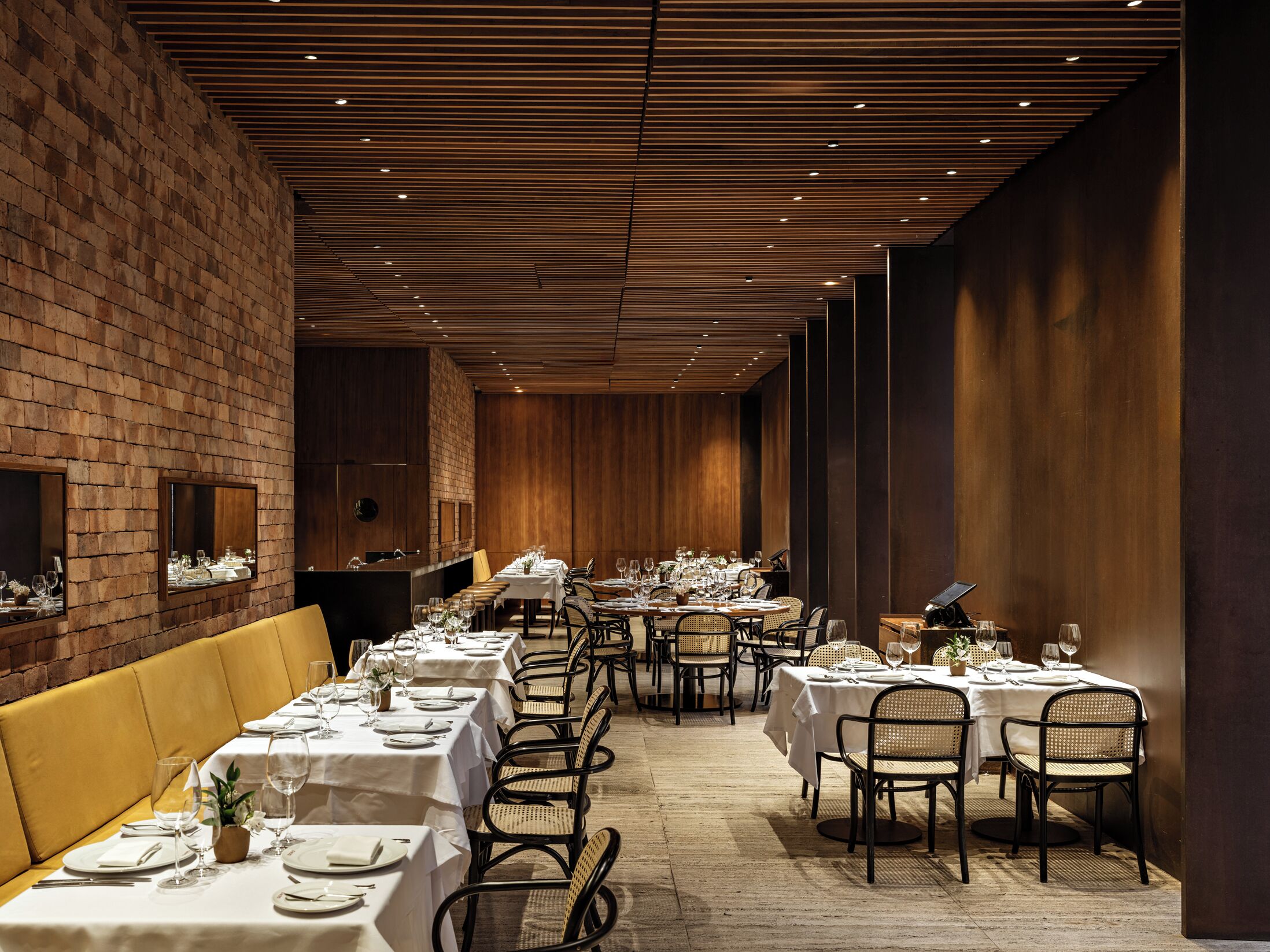 A long dining area with tables set for a meal, wooden paneling on the walls, and a brick accent wall.