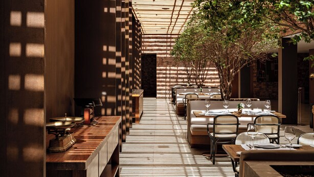 A restaurant with a modern interior, featuring wooden beams and a brick wall. Tables are set with wine glasses and plates, and there are chairs and benches for seating.