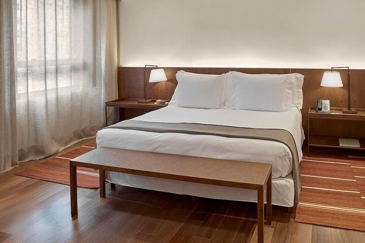 A hotel room with a large bed, two bedside tables, a bench, and a view of the city through the window.