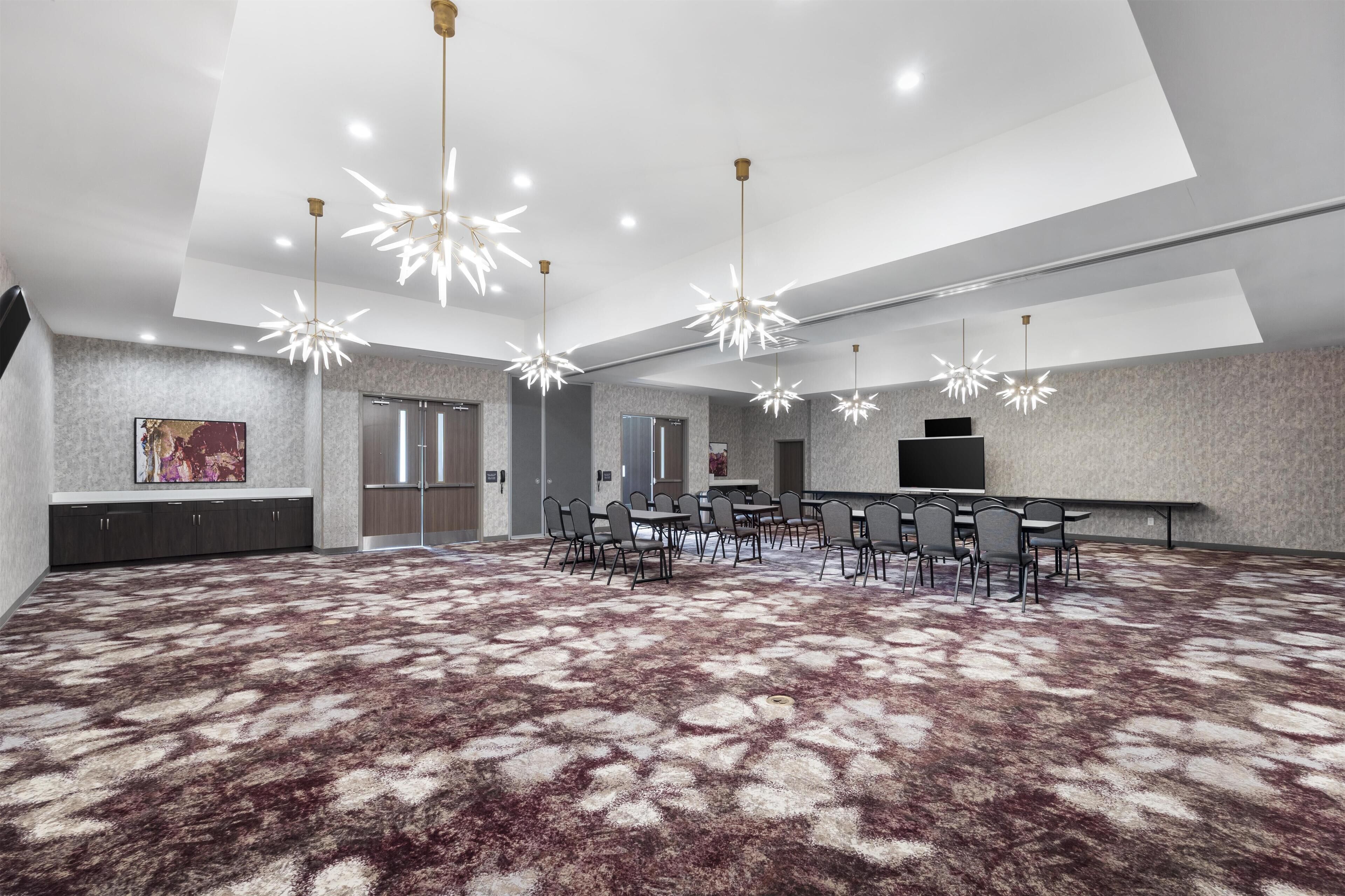 A spacious conference room with a patterned carpet, modern chandeliers, and a large flat-screen TV.