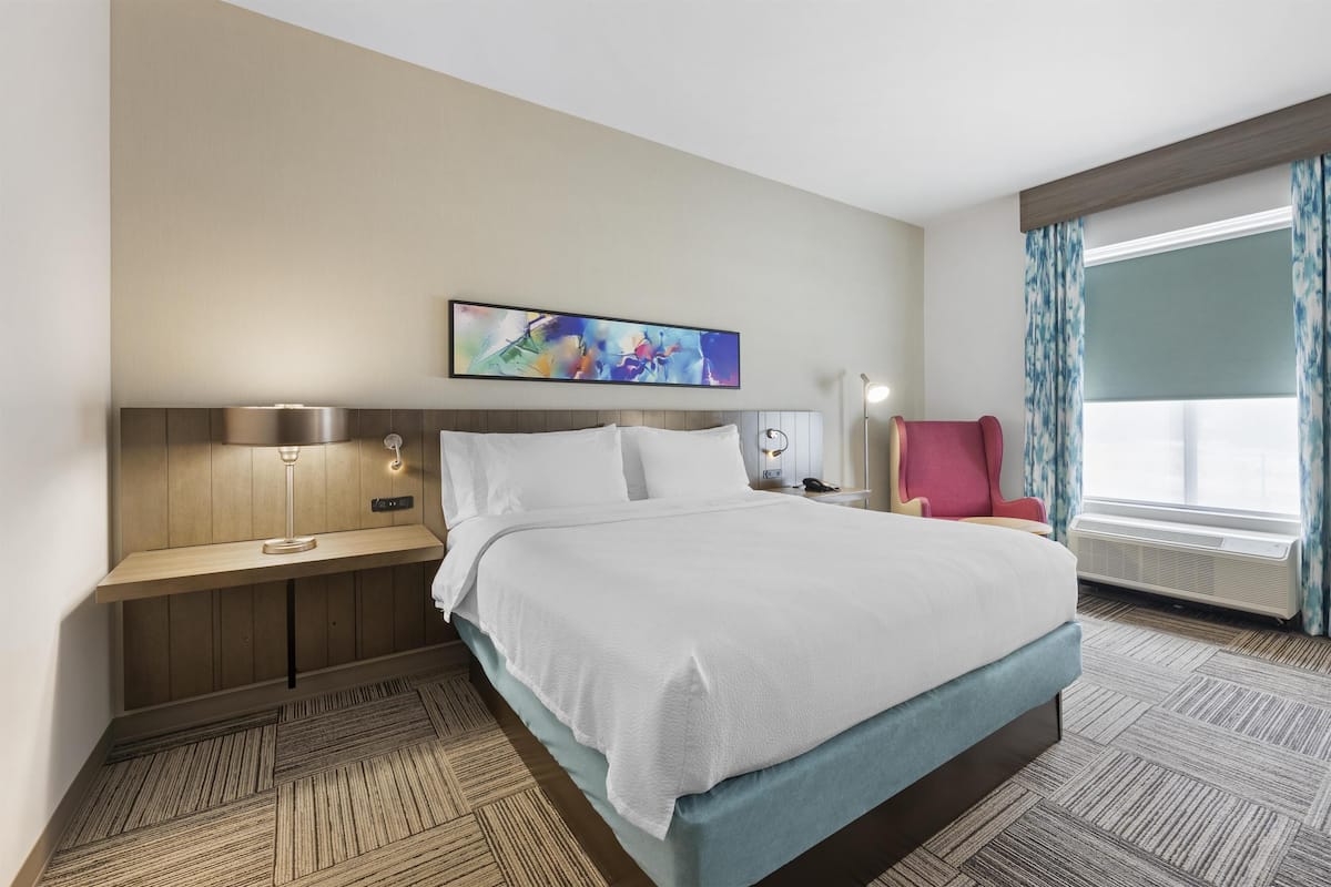 A hotel room with a bed, bedside table, lamp, and a window with curtains.