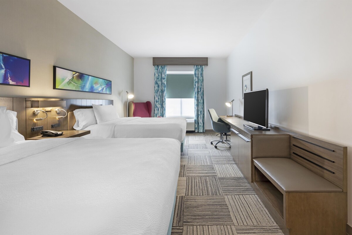 Habitación de hotel con dos camas, un escritorio, un televisor y obras de arte en las paredes.