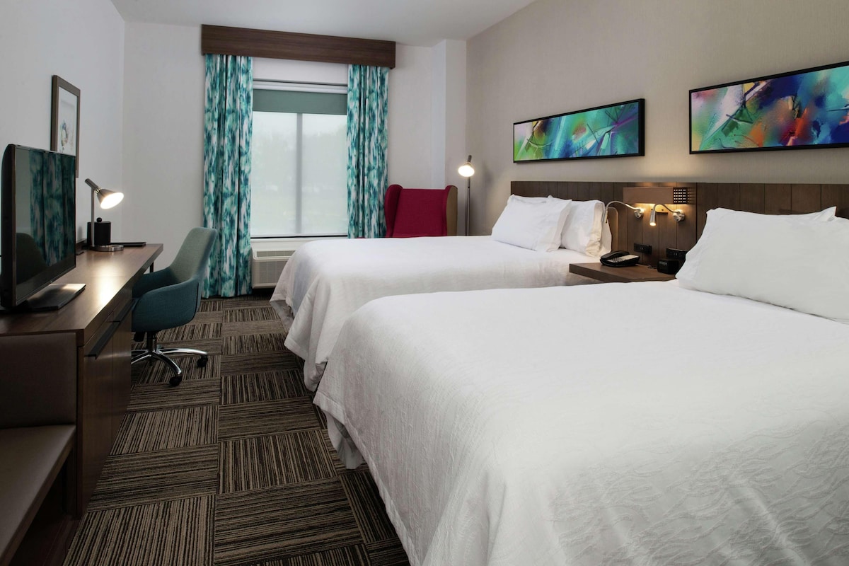 Quarto de hotel com duas camas, uma escrivaninha com computador, uma cadeira, televisão e obras de arte nas paredes.